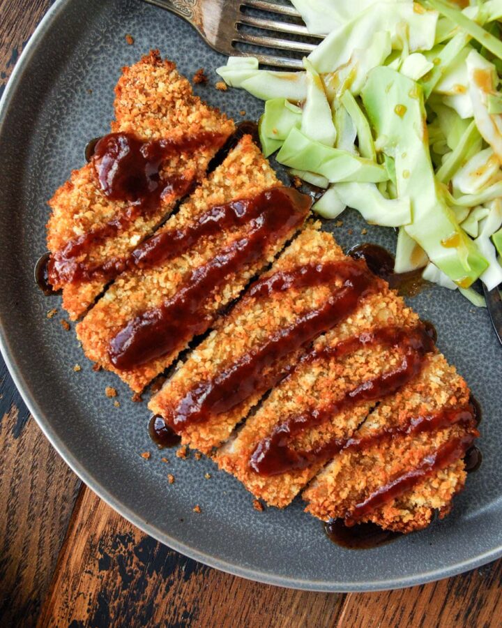Up close, above view of pork tonkatsu on a plate that has been sliced and has a drizzle of katsu sauce across the top and served with some shredded cabbage.