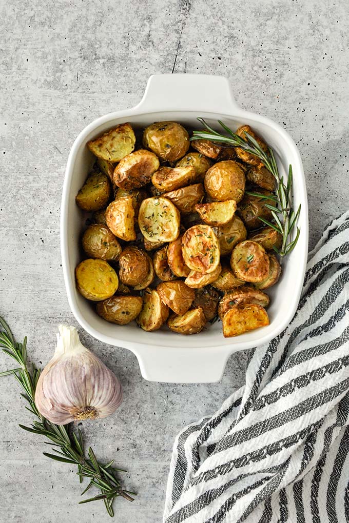 Above view of a baking dish of air fried potatoes with a striped towel, garlic bulb, and a sprig of fresh rosemary.