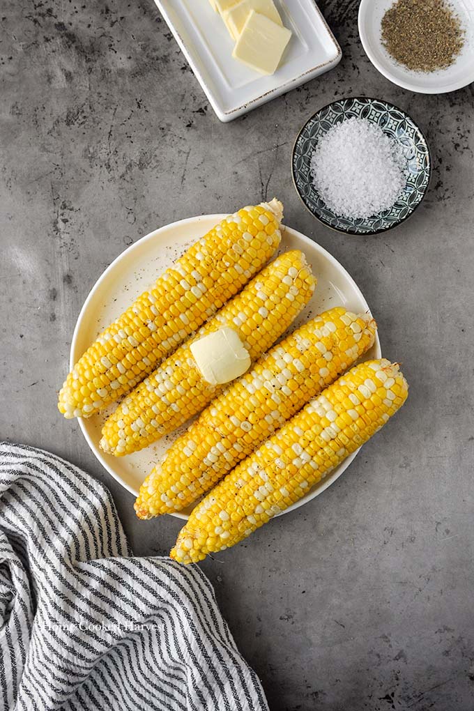 Above view of a plate of four cooked ears of corn with a striped towel, a bowl of salt and pepper, and a plate of butter slices.