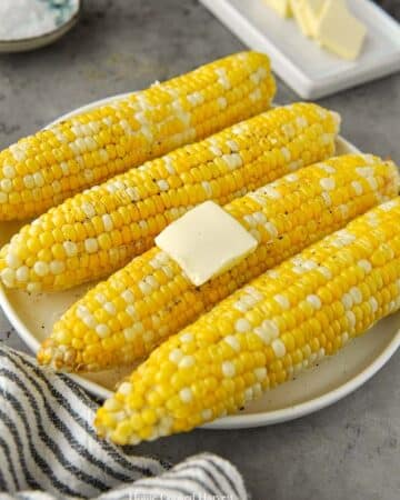 A plate of four cooked corn cobs with a pat of melty butter.