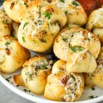 A garlic knot that has been dipped into marinara sauce and is being held above a plate of other buns.