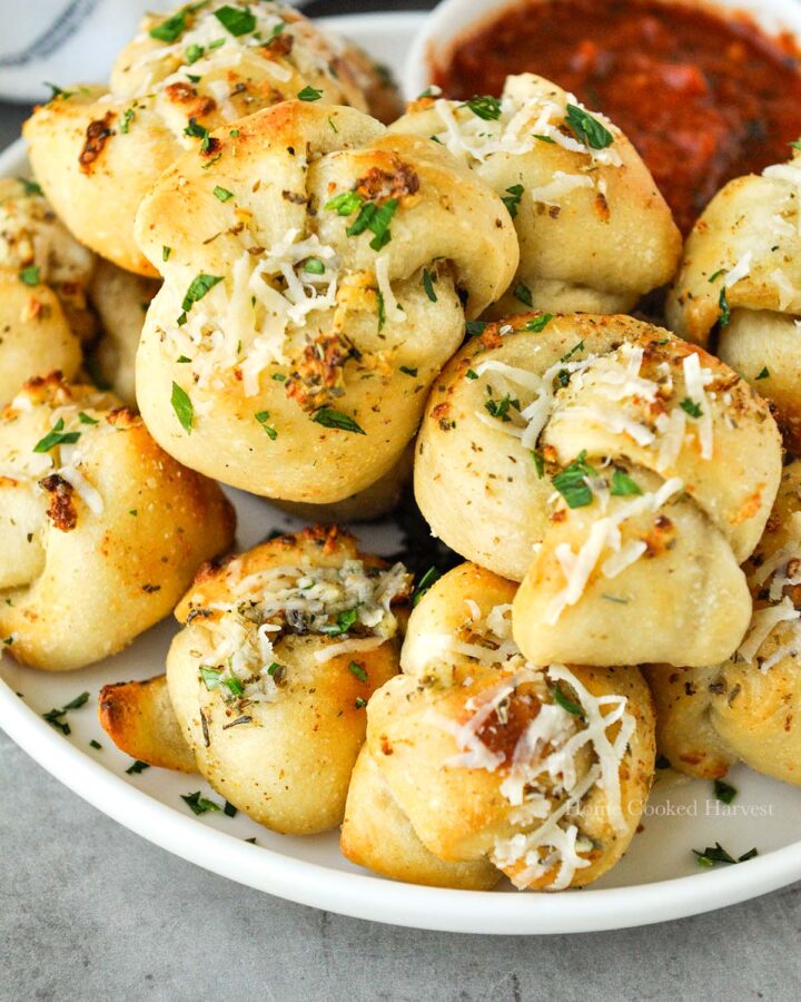 A garlic knot that has been dipped into marinara sauce and is being held above a plate of other buns.