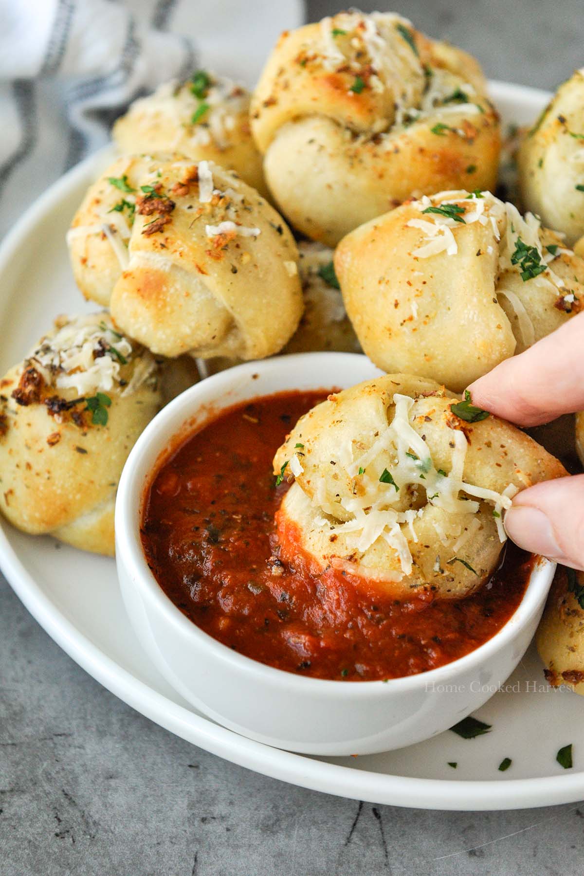 A garlic knot that is being dipped into marinara sauce with a plate of other buns.