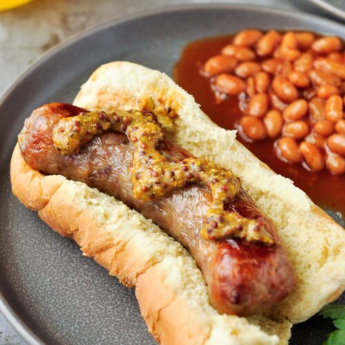Up close of a brat on a bun with mustard and a side of beans.
