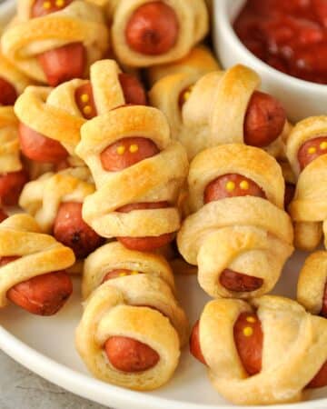 A plate of baked mini mummy's with a bowl of ketchup.