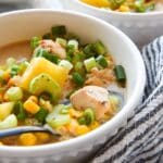 Up close bowl of chicken corn chowder with details of potatoes, chicken, corn, celery, and green onions. A striped towel is wrapped around the bowl and there is a second bowl of chowder in the background.