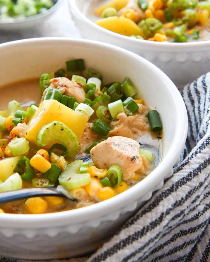 Up close bowl of chicken corn chowder with details of potatoes, chicken, corn, celery, and green onions. A striped towel is wrapped around the bowl and there is a second bowl of chowder in the background.