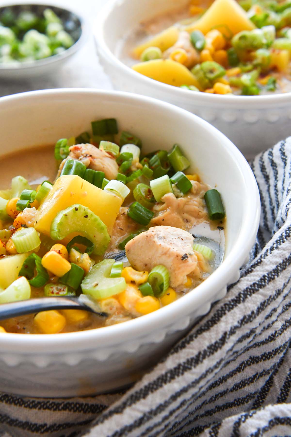 Up close bowl of chicken corn chowder with details of potatoes, chicken, corn, celery, and green onions. A striped towel is wrapped around the bowl and there is a second bowl of chowder in the background.