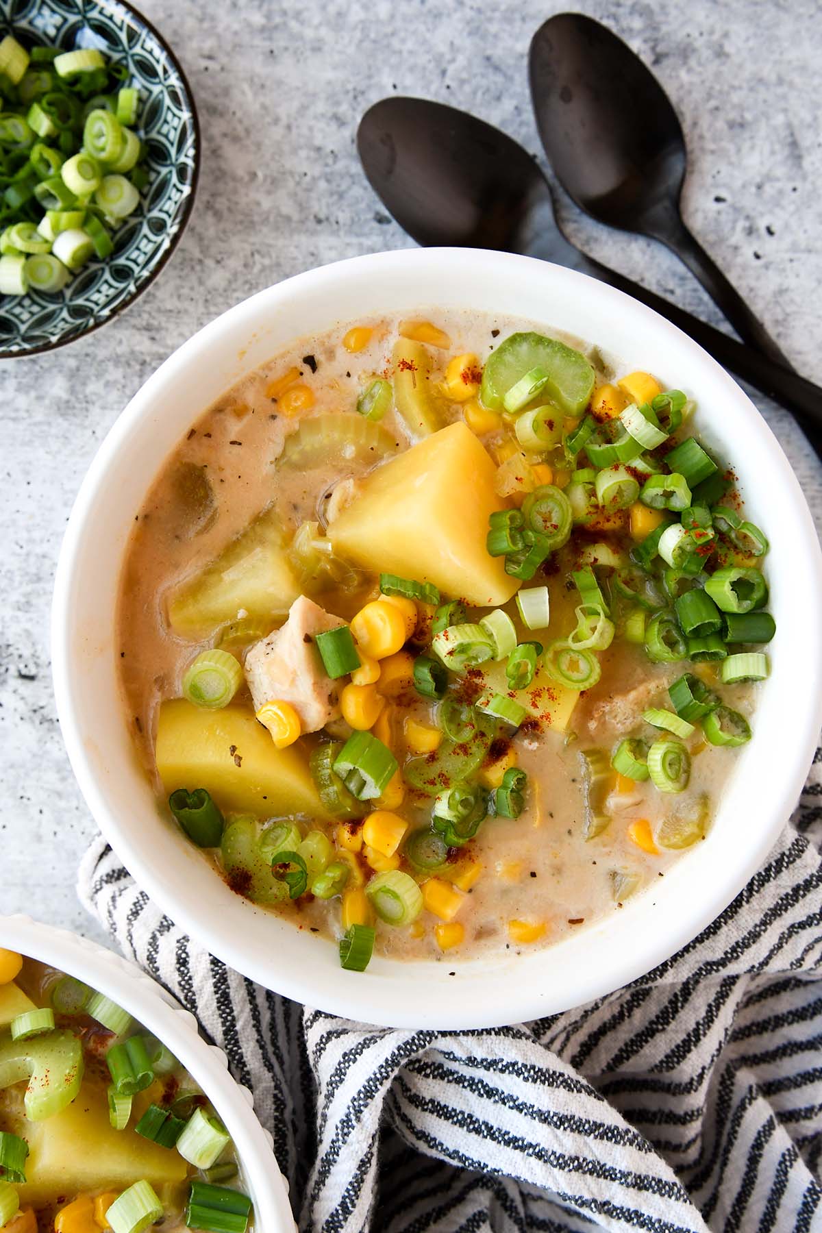 Above view of a bowl of chicken corn chowder with two black spoons, a striped towel is between that bowl of chowder and second bowl that isn't completely in the picture. 