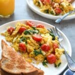 Two plates full of scrambled eggs with quinoa, tomatoes, and spinach with buttered toast and a glass of orange juice.