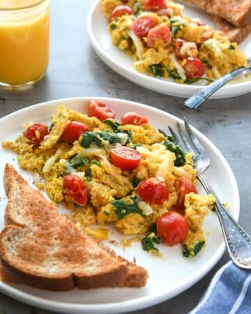 Two plates full of scrambled eggs with quinoa, tomatoes, and spinach with buttered toast and a glass of orange juice.