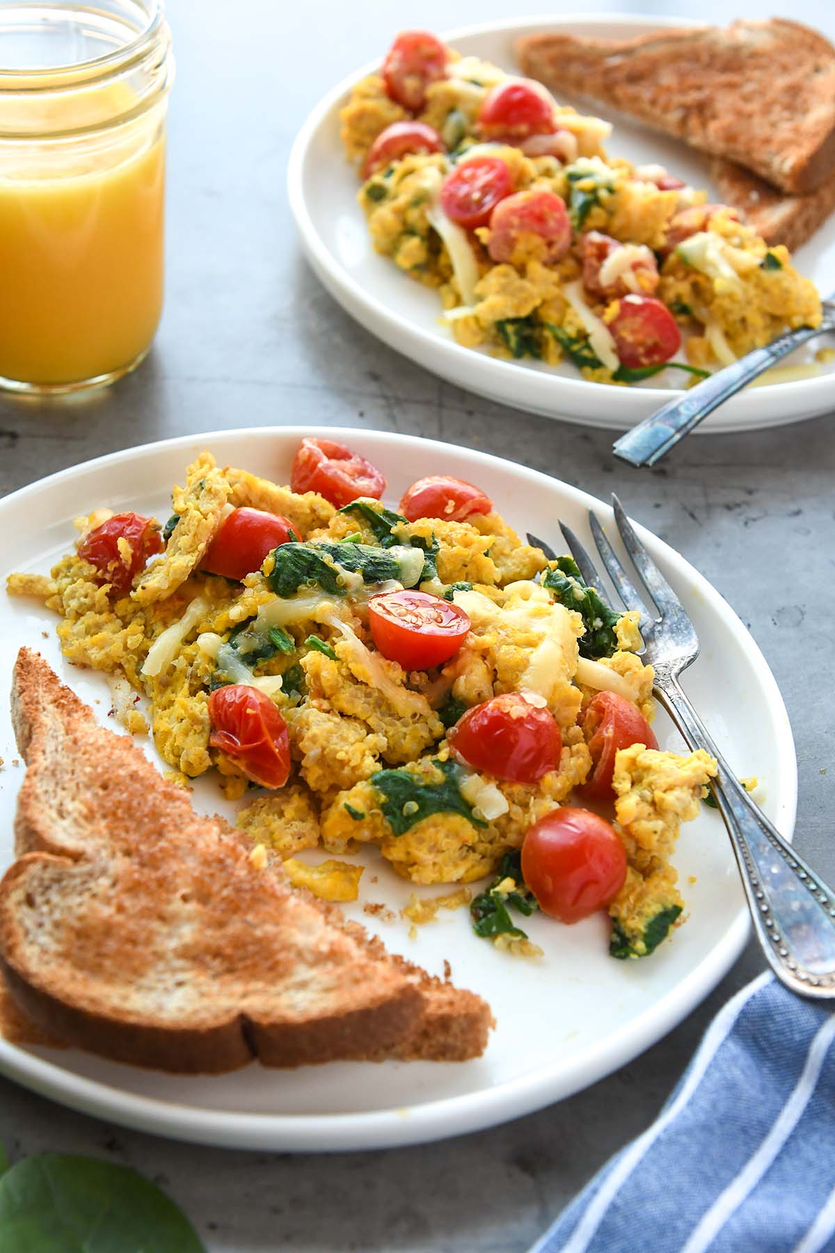 Two plates full of scrambled eggs with quinoa, tomatoes, and spinach with buttered toast and a glass of orange juice.