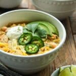 Two big bowls of chili topped with sour cream, avocado, cilantro, and jalapeno slices. A small bowl of lime wedges are in front of the bowls with a striped towel and a small bowl of jalapeños in the background.