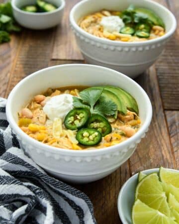 Two bowls of chicken chili with some lime slices and a bowl of jalapeno slices.