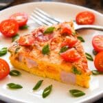 Up close of a slice of frittata on a plate sprinkled with cut tomatoes and green onions.