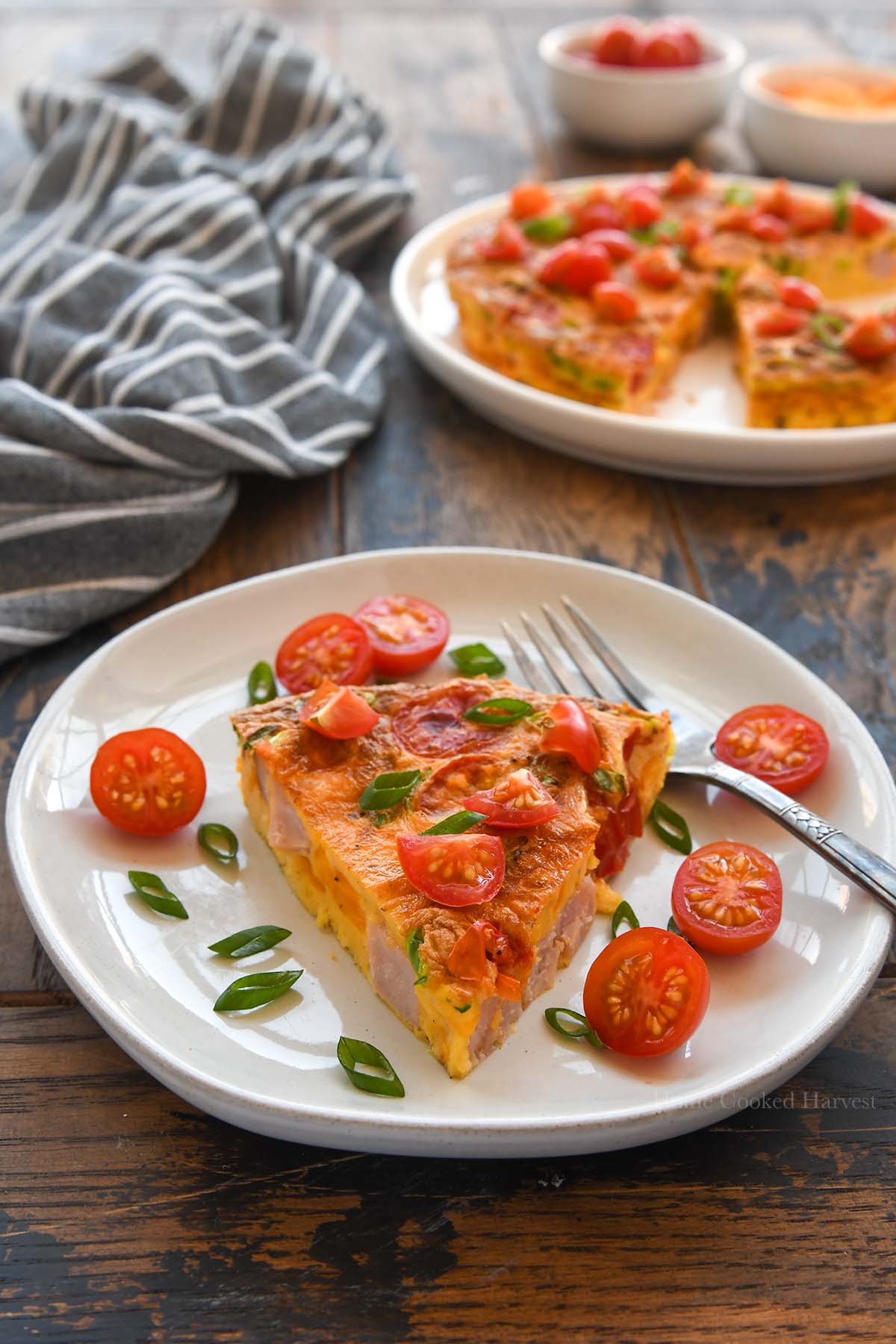 A plate of a slice of frittata decorated with sliced green onions and tomatoes. A plate of the rest of the frittata in the background.