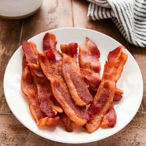 A white plate full of cooked bacon with a striped towel and eggs in a bowl.