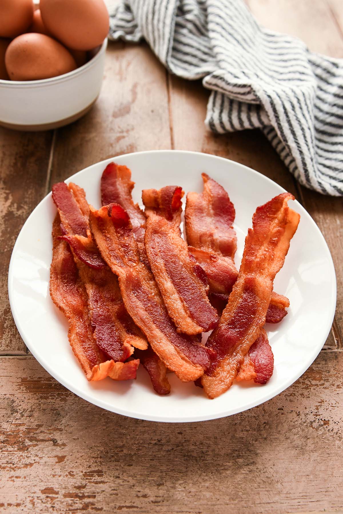 A white plate full of cooked bacon with a striped towel and eggs in a bowl.