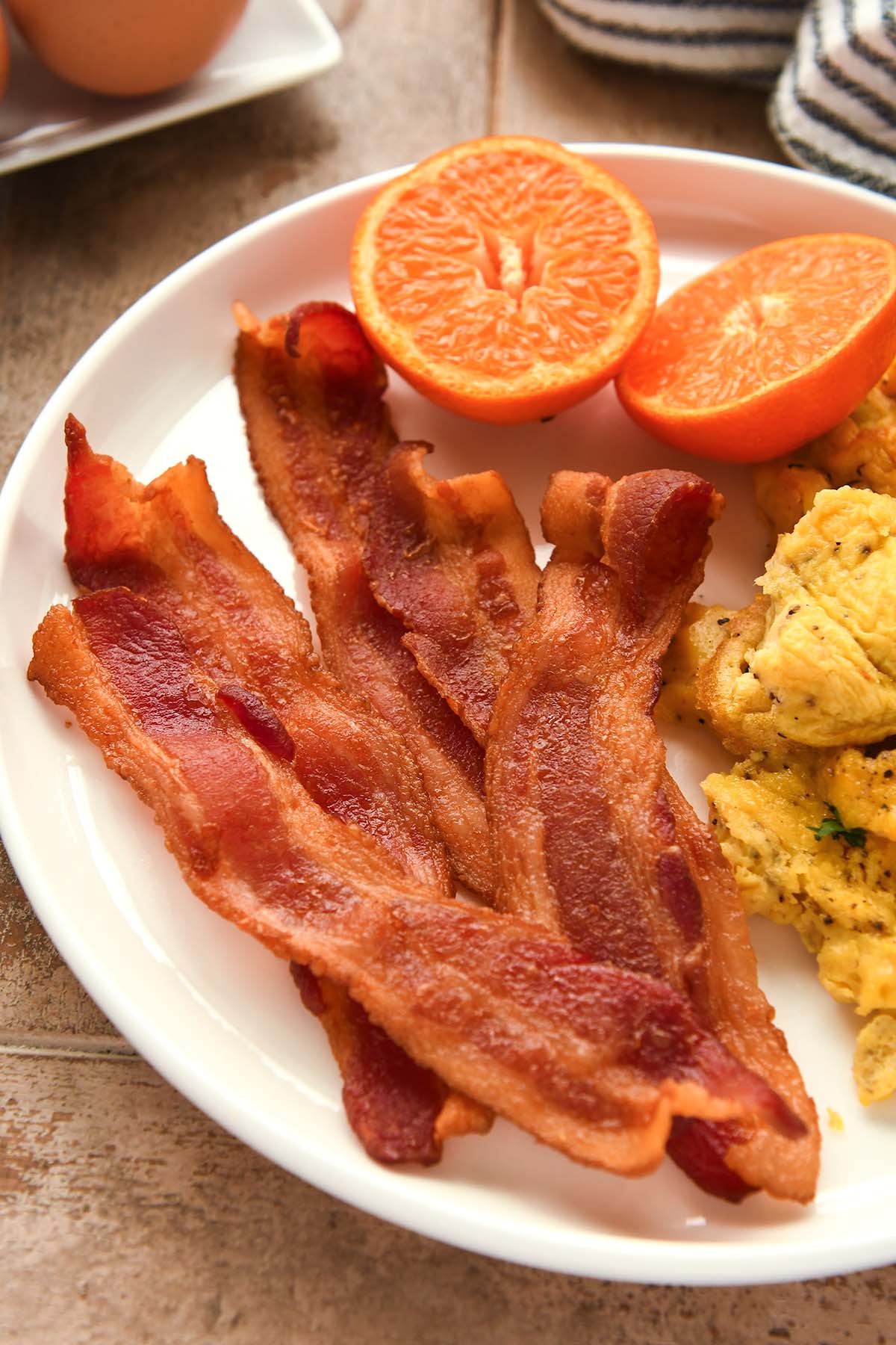 A white plate with a couple strips of cooked bacon, a cut orange, and some scrambled eggs.