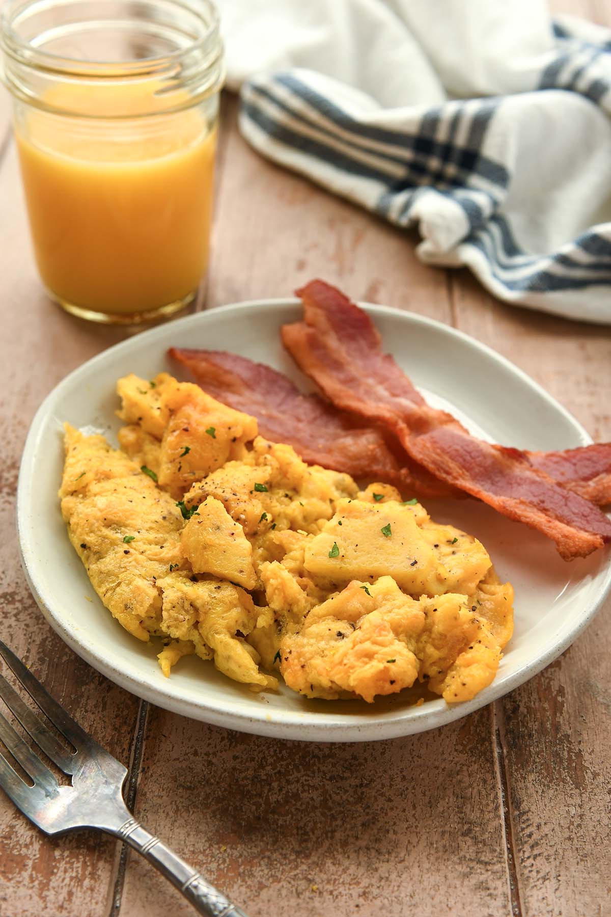 A plate with air fryer scrambled eggs and some bacon. A glass of orange juice and a striped towel are in the background.