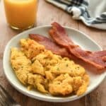 A plate with air fryer scrambled eggs and some bacon. A glass of orange juice and a striped towel are in the background.