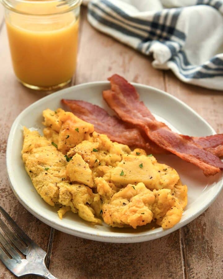 A plate with air fryer scrambled eggs and some bacon. A glass of orange juice and a striped towel are in the background.