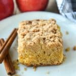 A slice of apple crumble cake on a white plate with two cinnamon sticks. Apples and a towel in the background.