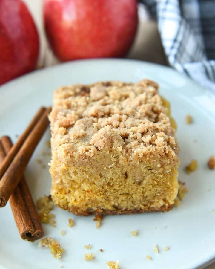 A slice of apple crumble cake on a white plate with two cinnamon sticks. Apples and a towel in the background.