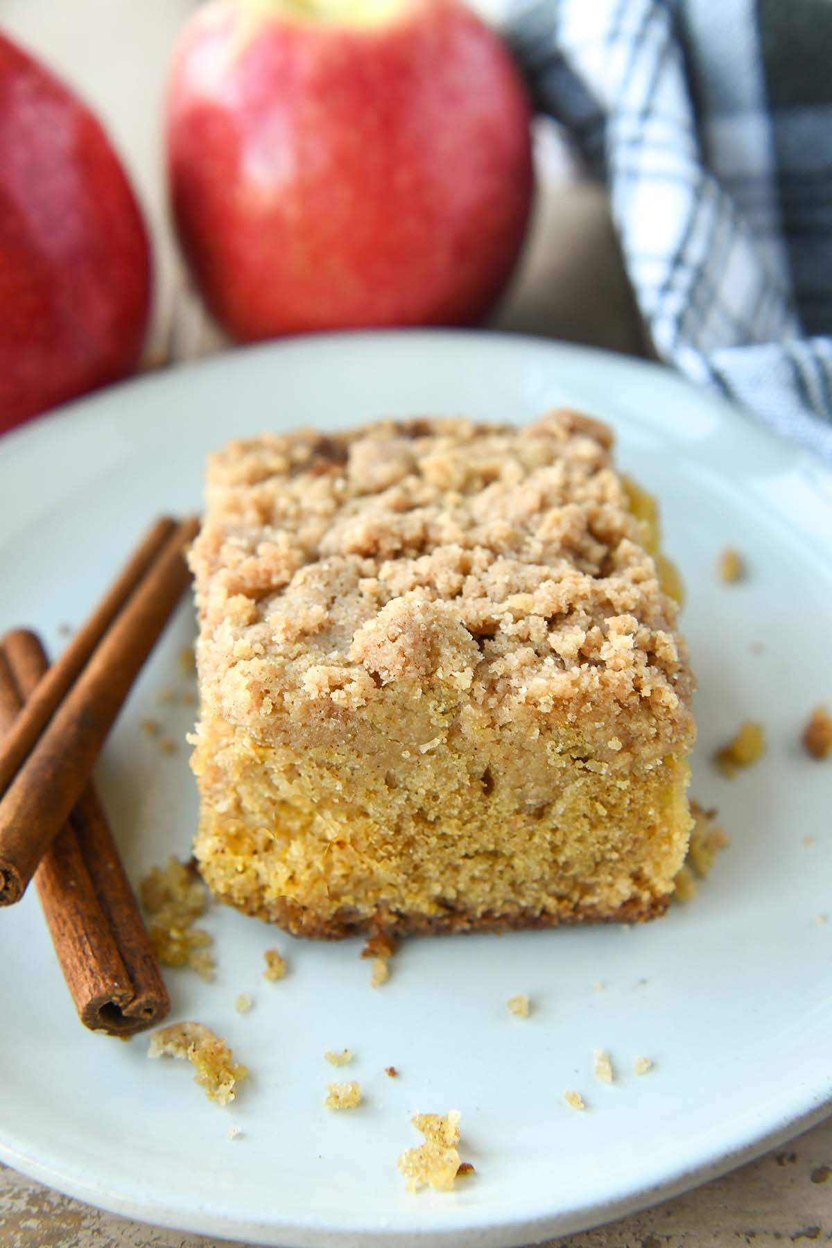 A slice of apple crumble cake on a white plate with two cinnamon sticks. Apples and a towel in the background.