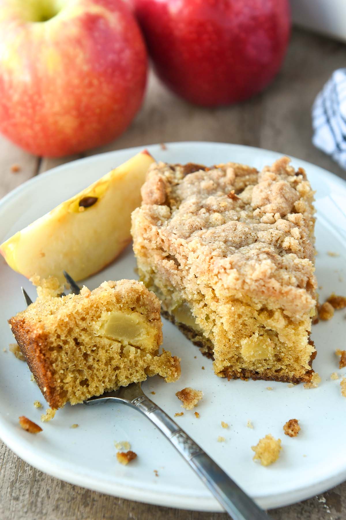 A forkful of apple cake next to a piece of cake with an apple slice. Apples and a striped towel in the background.