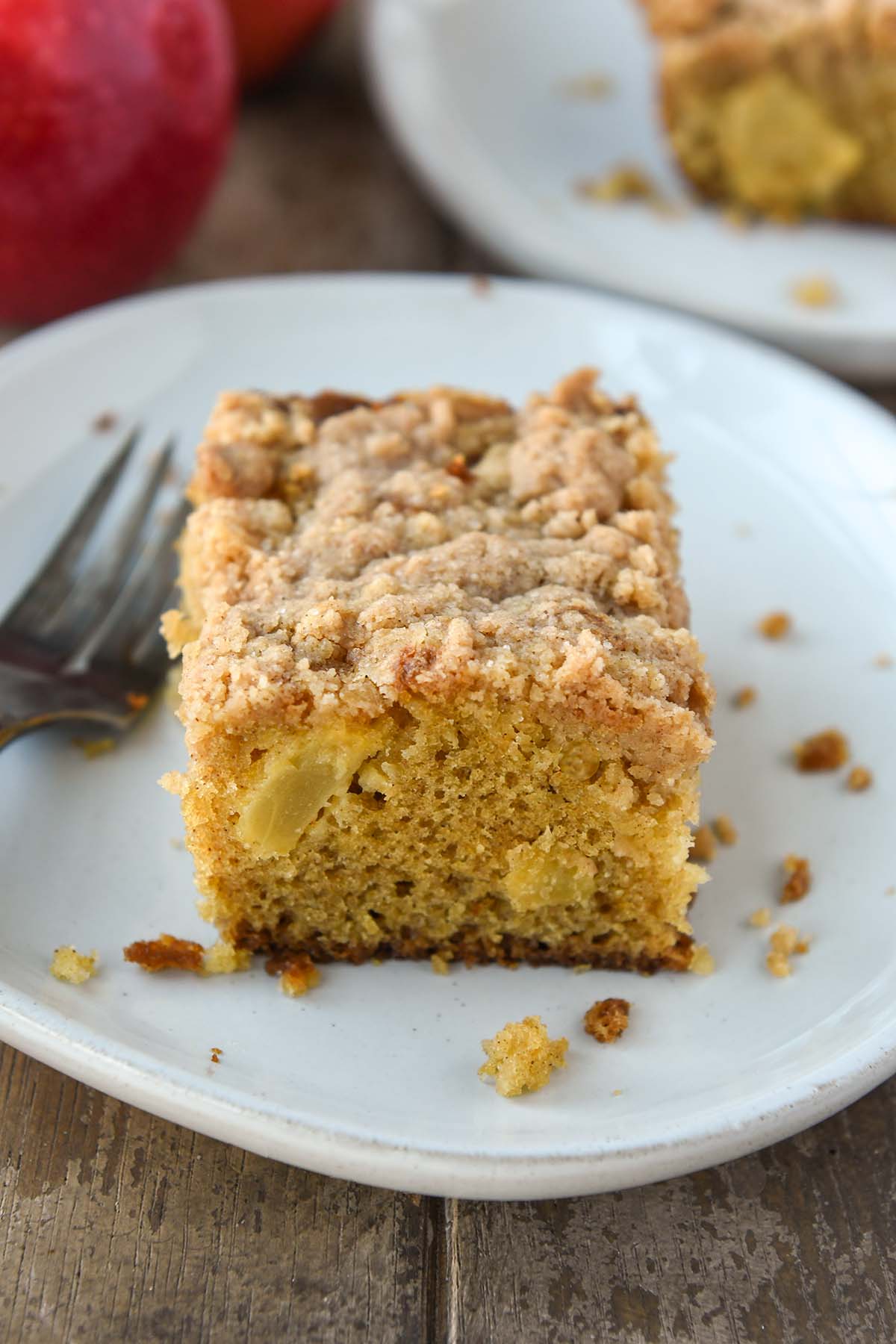 A slice of apple crumble cake on a white plate with two cinnamon sticks. Apples and a towel in the background.