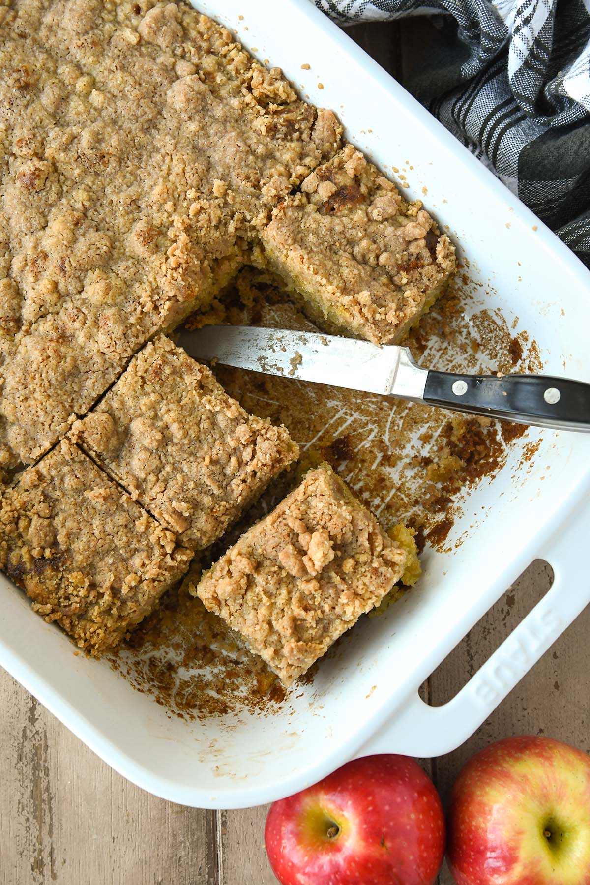 Above the pan of cooked apple cake with slices cut and some removed. A knife rests in the pan. Two apples and a towel are next to the pan.