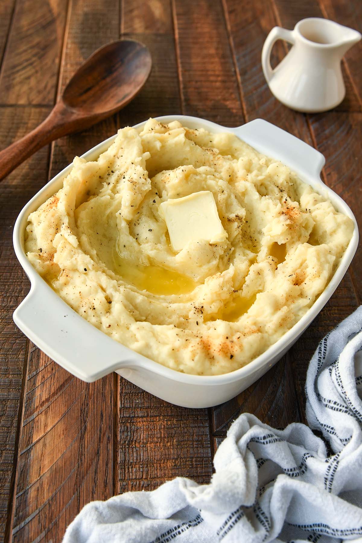 An above view of a square dish filled with mashed potatoes swirled with butter and a sprinkling pepper and seasoned salt. With a white and black striped towel, small pitcher of gravy and a wooden spoon.
