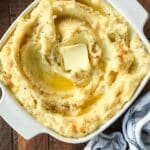 An above view of a square dish filled with mashed potatoes swirled with butter and a sprinkling pepper and seasoned salt. With a white and black striped towel.