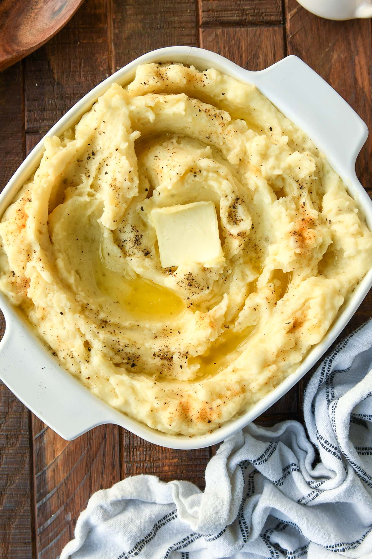 An above view of a square dish filled with mashed potatoes swirled with butter and a sprinkling pepper and seasoned salt. With a white and black striped towel.
