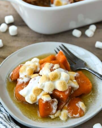 A plateful candied yams with a fork and a baking dish and marshmallows around.