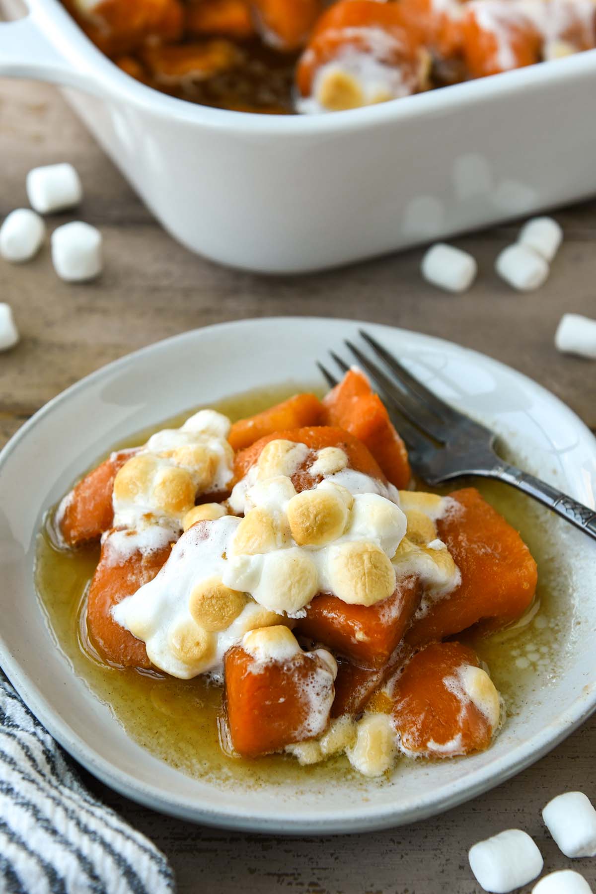 A plateful candied yams with a fork and a baking dish and marshmallows around.