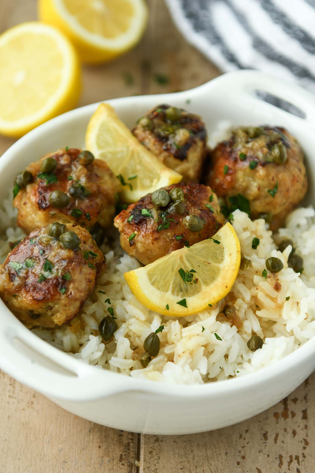 A white bowl of meatballs over rice with some lemon slices and a striped towel.