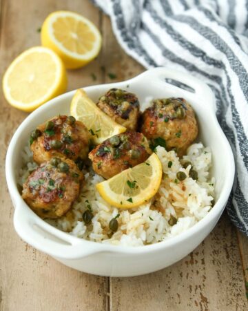 A white bowl of meatballs over rice with some lemon slices and a striped towel.