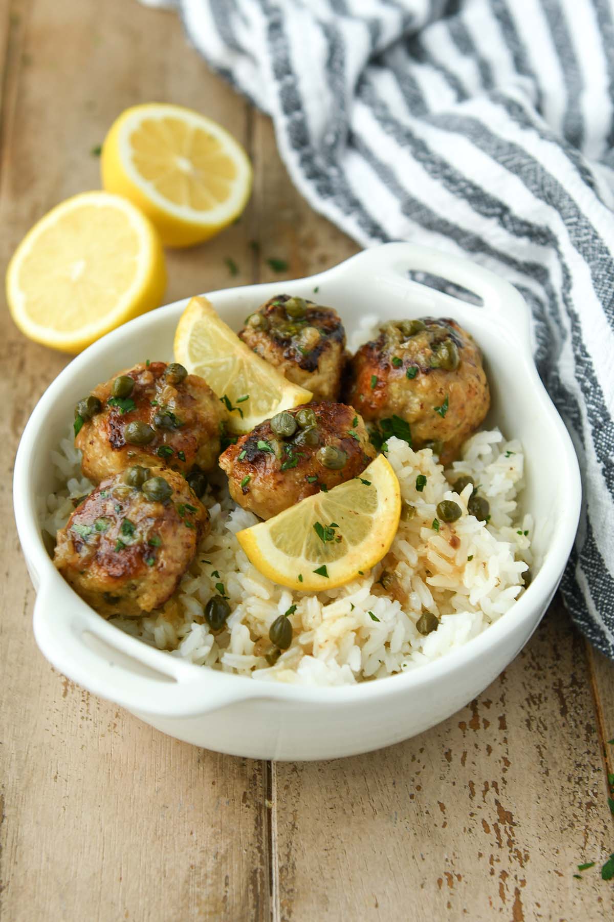 A white bowl of meatballs over rice with some lemon slices and a striped towel.