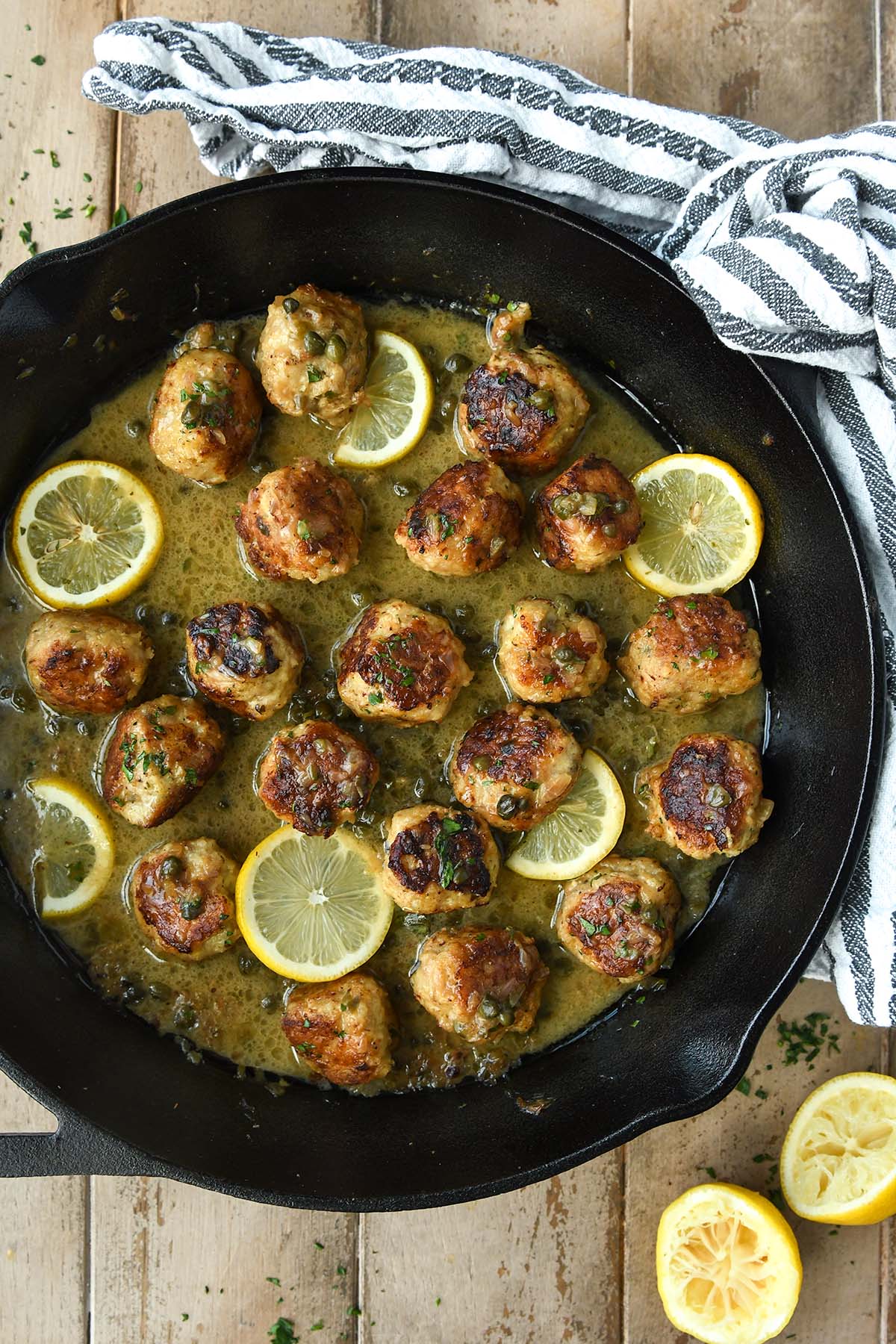Above view of a black skillet filled with baked meatballs with lemon slices and a striped towel in the background.