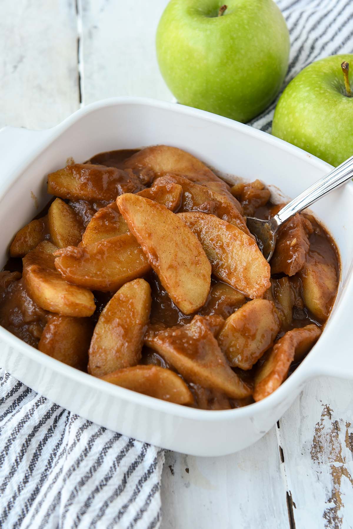 A square dish of the cooked apples with a spoon. Two green apples and a striped towel in the background.