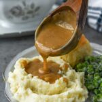 A ladle of gravy being poured onto mashed potatoes with a roll and peas.