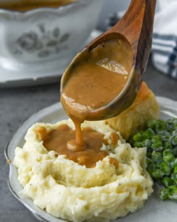 A ladle of gravy being poured onto mashed potatoes with a roll and peas.
