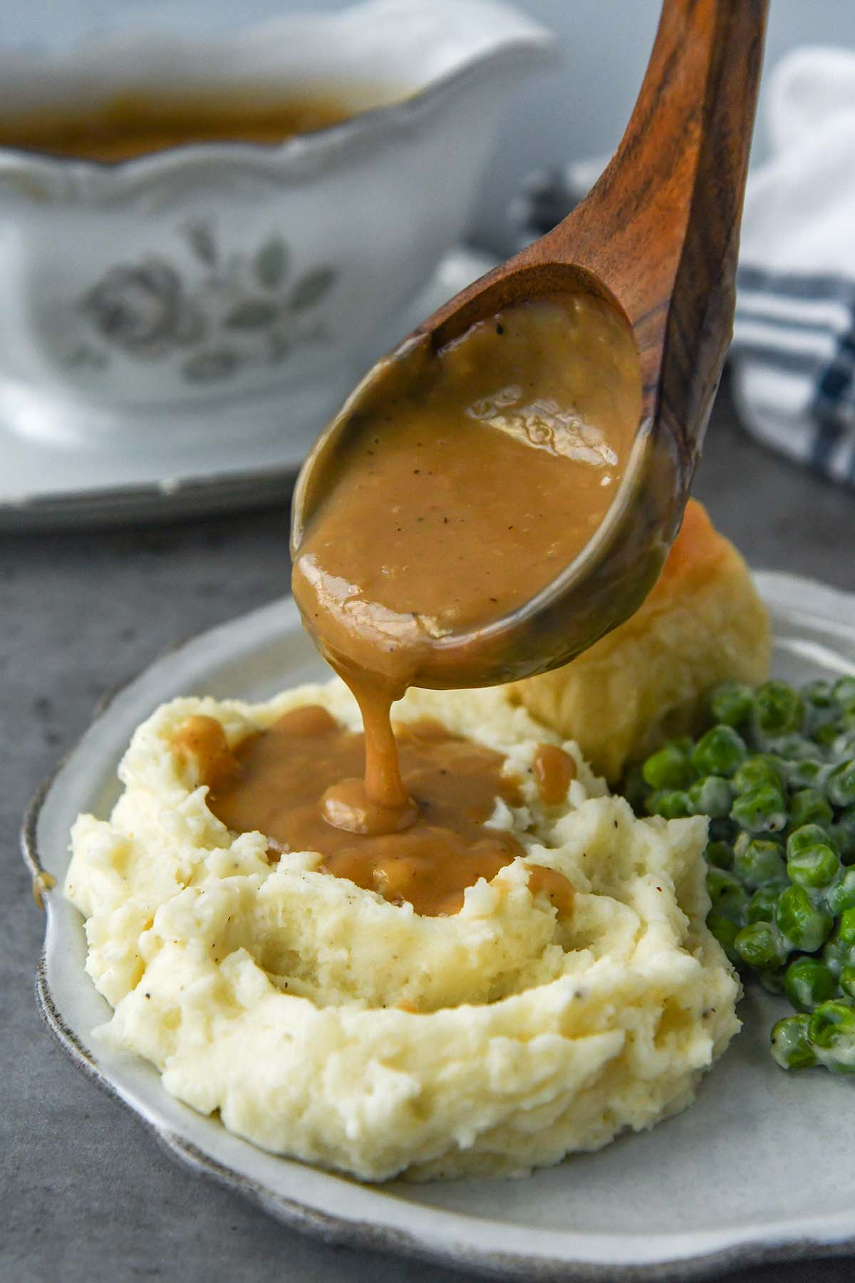 A ladle of gravy being poured onto mashed potatoes with a roll and peas.
