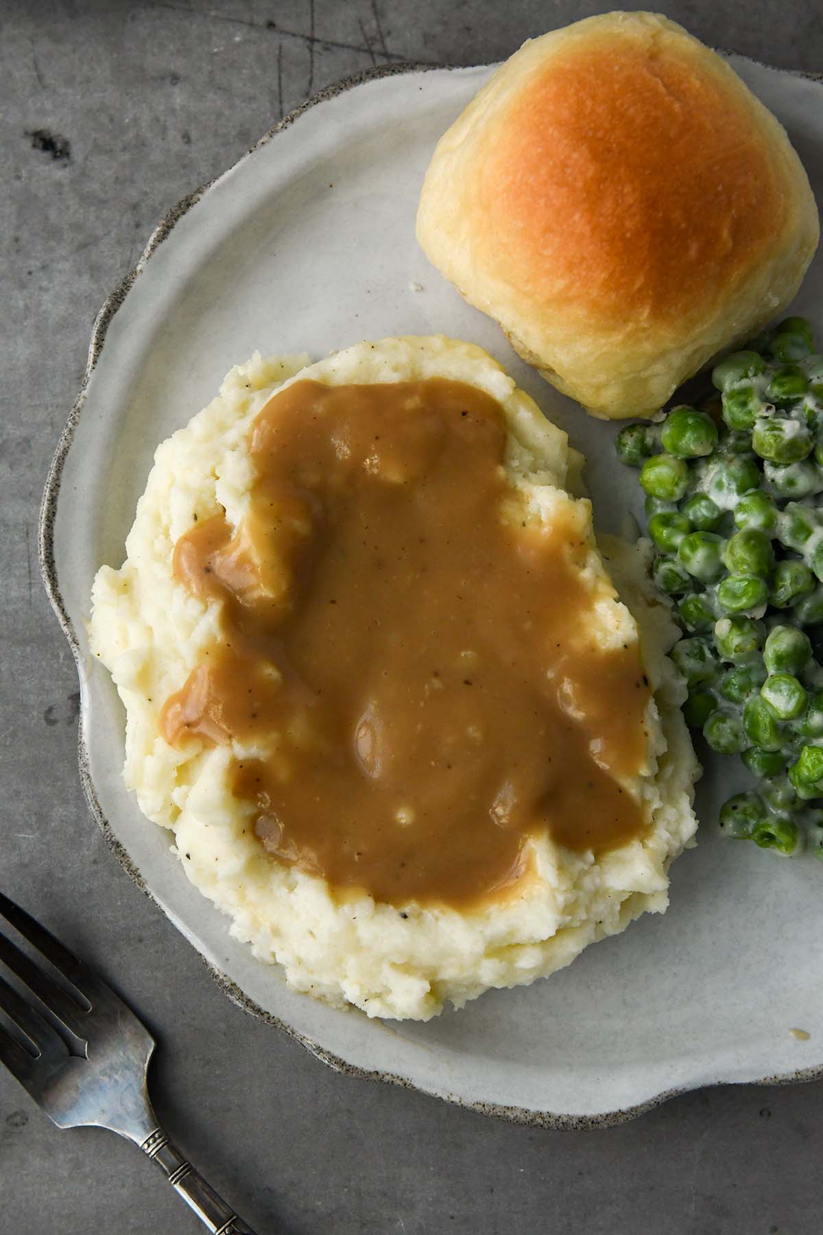 Above view of a plate of gravy coated mashed potatoes, a roll, and peas.