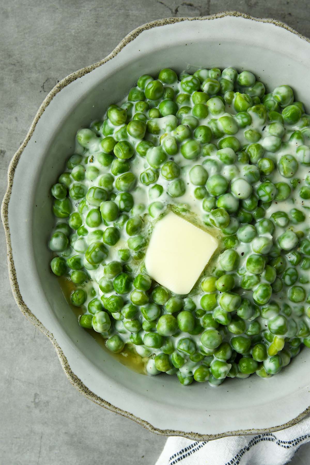 An above view of a bowl of creamed peas with a melty pat of butter and a white and black striped towel.