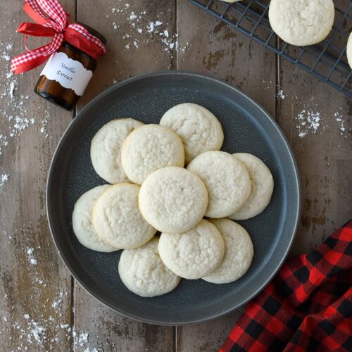 An above view of meltaway cookies on a gray plate with a bottle of vanilla extract, a cooling rack with more cookies, and a plaid towel.