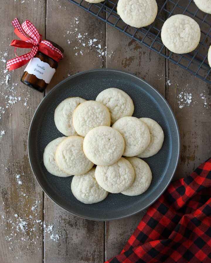 An above view of meltaway cookies on a gray plate with a bottle of vanilla extract, a cooling rack with more cookies, and a plaid towel.
