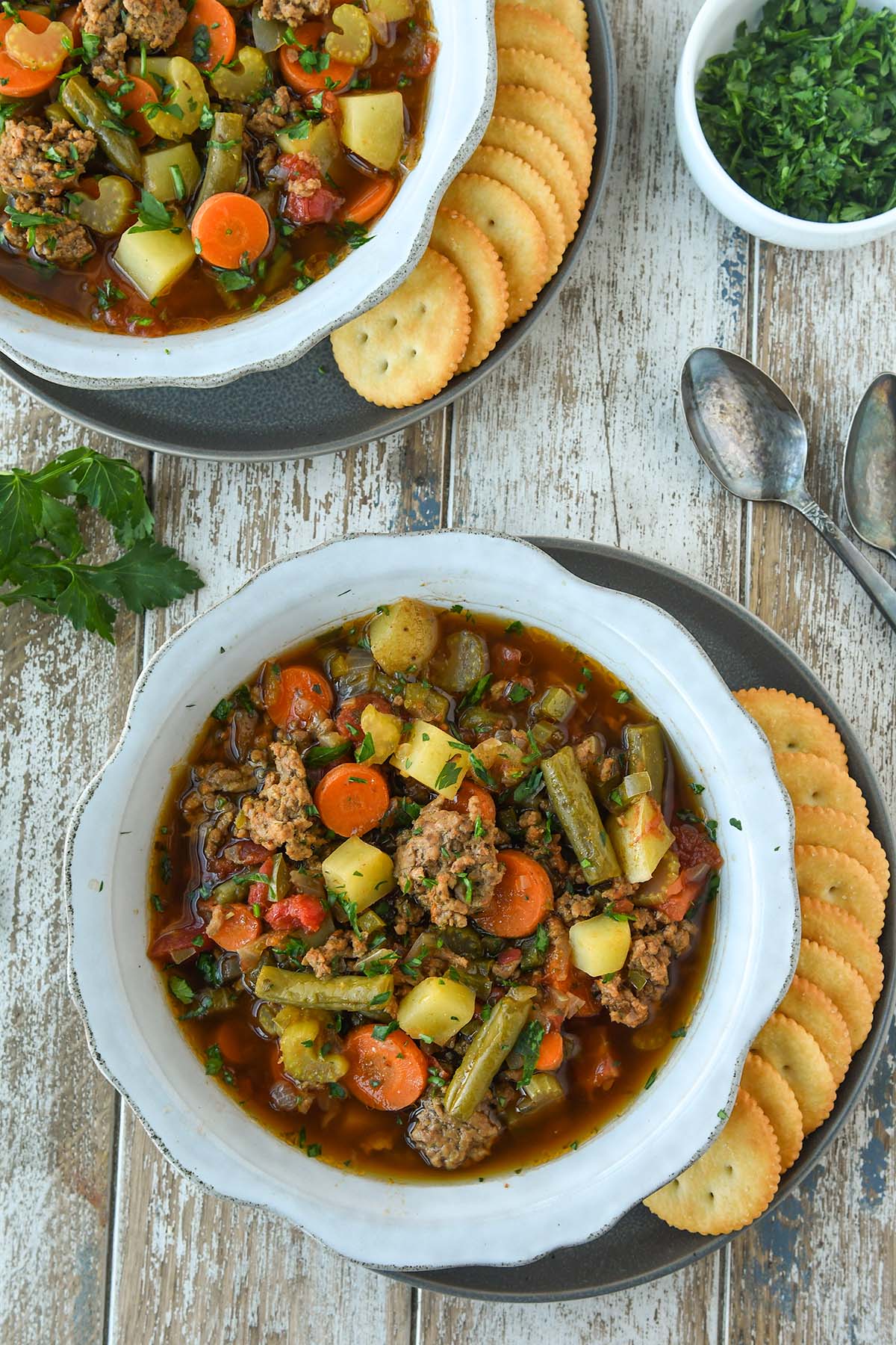 Above view of two bowls of hamburger soup with ritz crackers and spoons.